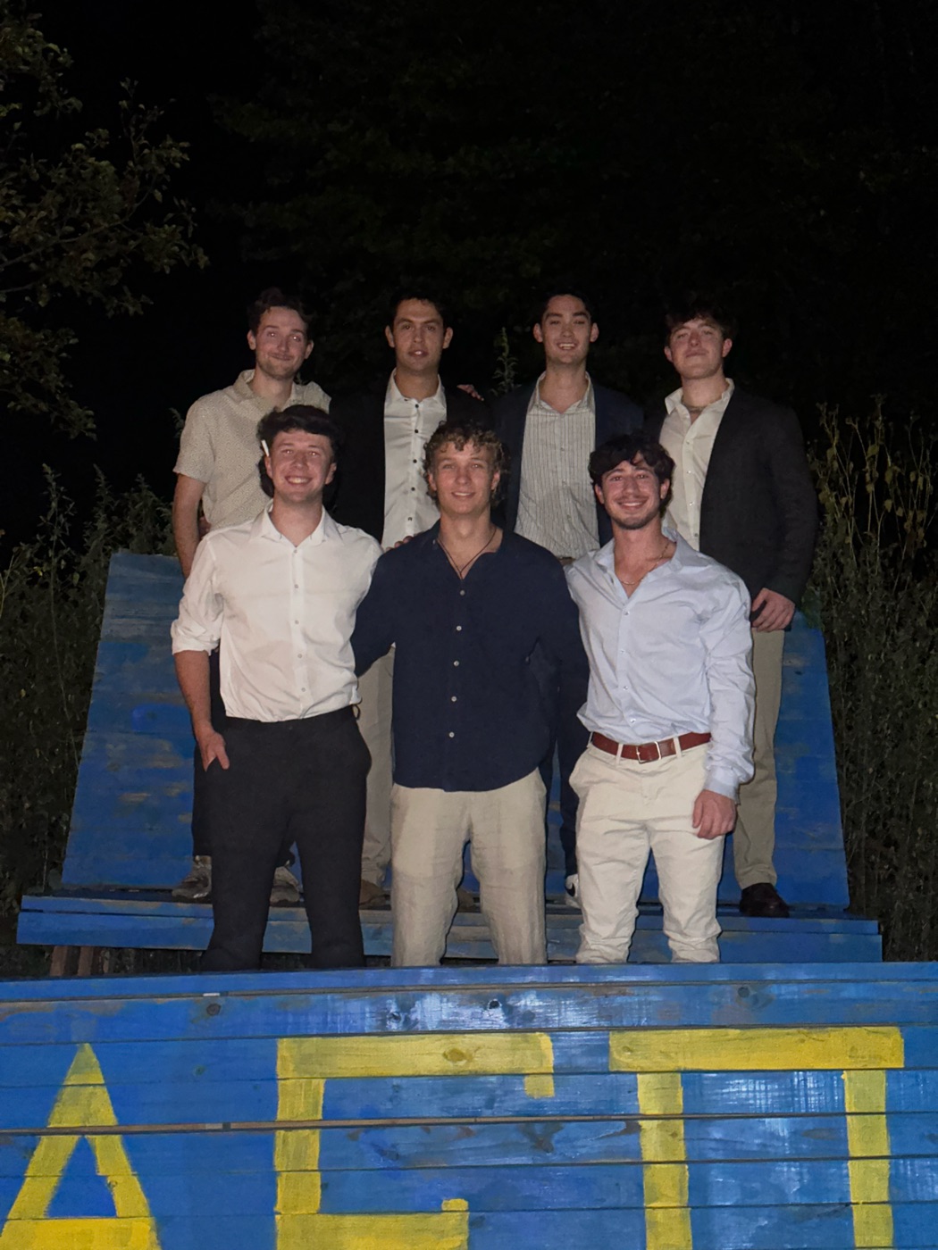 Kevin with six Alpha Epsilon Pi brothers at night, standing on a painted wooden platform with the chapter's letters
