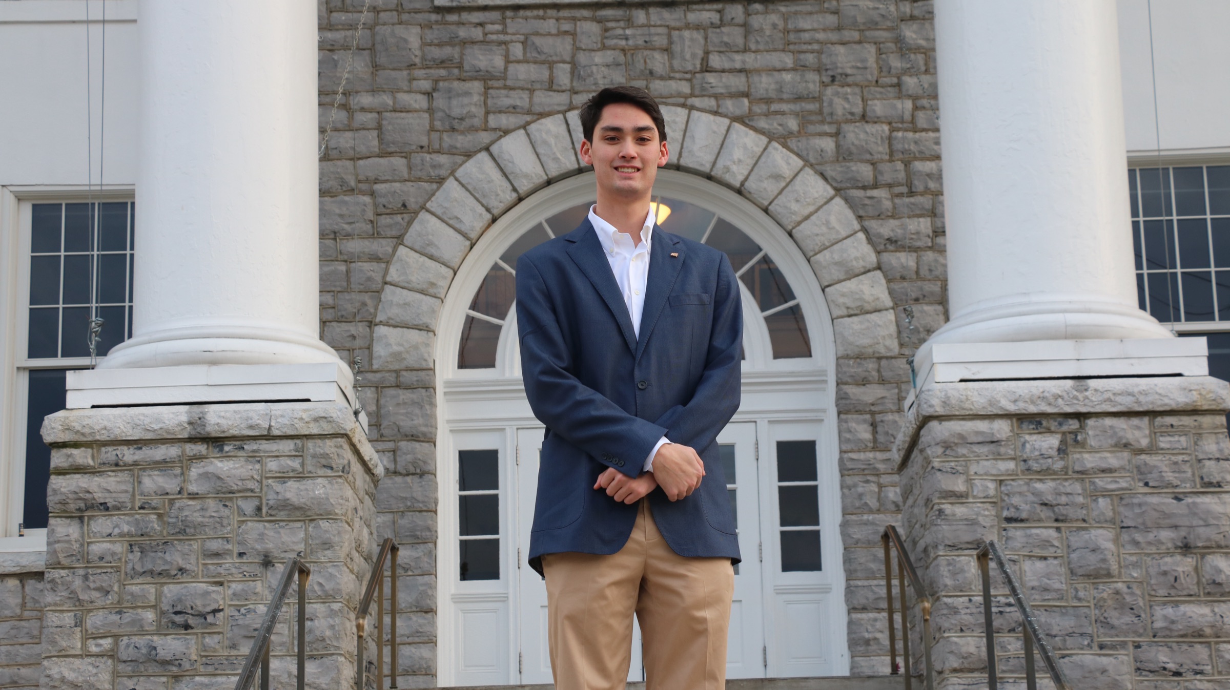 Kevin Backer in a navy blazer and khakis, standing on the bluestone steps of a JMU building between two white columns